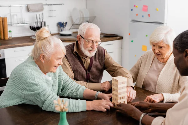 Веселі міжрасові пенсіонери посміхаються, граючи у дерев'яні блоки вежі вдома — стокове фото