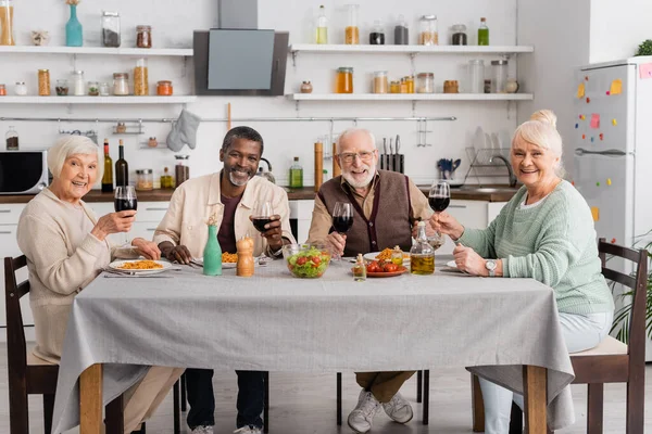 Щасливі міжрасові пенсіонери тримають окуляри з червоним вином біля смачного обіду на столі — стокове фото