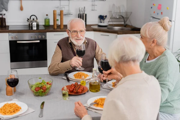 Happy senior man clinking glasses of red wine with retired women near tasty lunch on table — Stock Photo