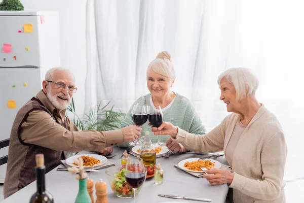 Felices amigos mayores tintineo vasos con vino tinto cerca de sabroso almuerzo en la mesa - foto de stock