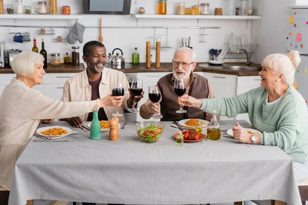 Счастливые межрасовые пенсионеры звон стаканов с красным вином рядом вкусный обед на столе — стоковое фото