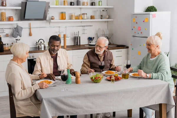 Feliz interracial e idosos amigos tendo delicioso almoço perto de copos de vinho tinto — Fotografia de Stock