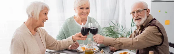 Felices amigos mayores tintineo vasos con vino tinto cerca de sabroso almuerzo en la mesa, pancarta - foto de stock