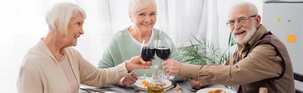 Happy senior friends clinking glasses with red wine near tasty lunch on table, banner — Stock Photo