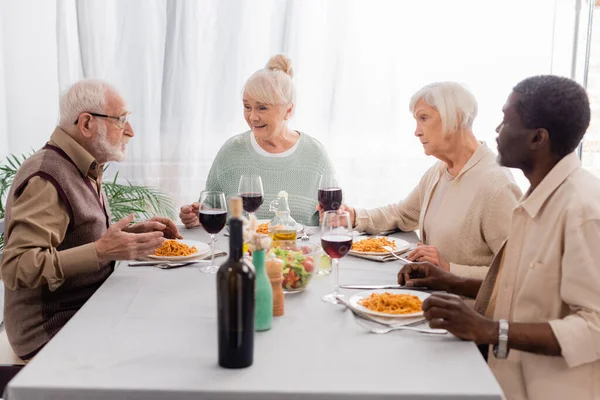Heureux amis multiculturels et aînés parler tout en ayant délicieux déjeuner près de verres de vin rouge — Photo de stock