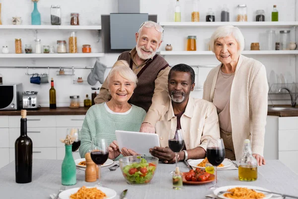 Personas mayores multiculturales sosteniendo tableta digital cerca de amigos felices y sabrosa comida en la mesa - foto de stock