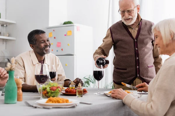 Homem sênior em óculos derramando vinho tinto em vidro perto de amigos inter-raciais felizes — Fotografia de Stock