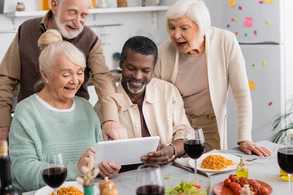 Personas jubiladas multiculturales mirando tableta digital con amigos mayores felices cerca de la comida sabrosa en la mesa - foto de stock