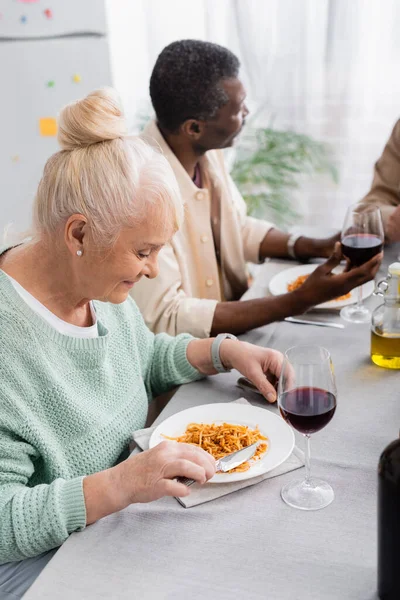Mujer mayor feliz mirando el plato con pasta cerca de amigo afroamericano durante el almuerzo - foto de stock