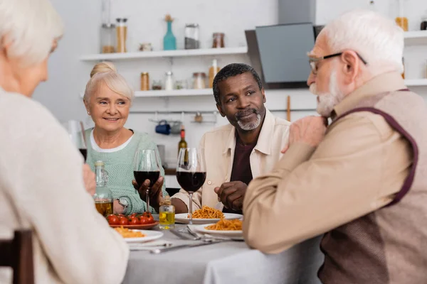 Pessoas multiculturais e seniores felizes olhando para o homem aposentado, tendo delicioso almoço juntos — Fotografia de Stock