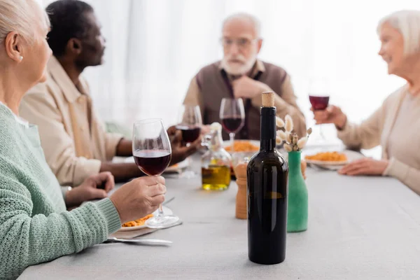 Botella de vino tinto cerca de amigos multiculturales y senior teniendo un delicioso almuerzo juntos en un fondo borroso - foto de stock