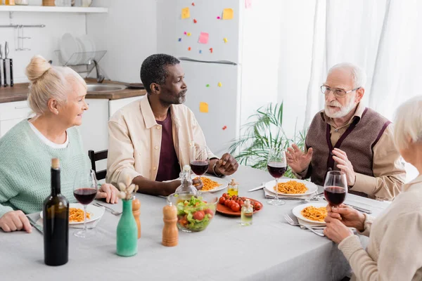 Happy multicultural pensioners looking at senior man during lunch — Stock Photo