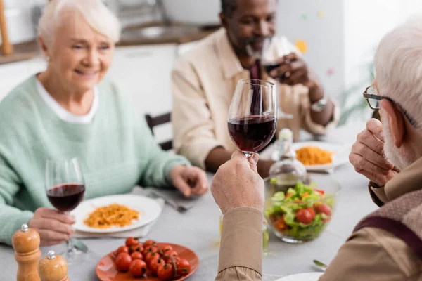 Senior man holding glass of red wine near multicultural friends on blurred background — Stock Photo