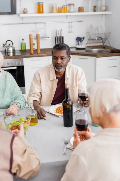 Senior afrikanisch-amerikanischer Mann hält ein Glas Rotwein in der Nähe pensionierter Freunde auf verschwommenem Vordergrund — Stockfoto