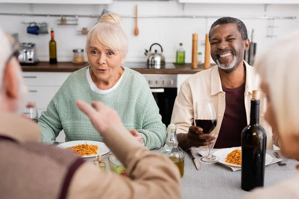 Amazed senior woman looking at man gesturing near interracial friends while having lunch together — Stock Photo