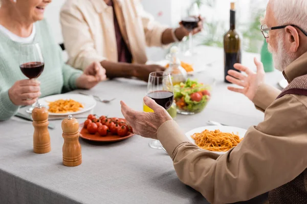 Hombre mayor en gafas gestos mientras almorza con amigos multiculturales - foto de stock