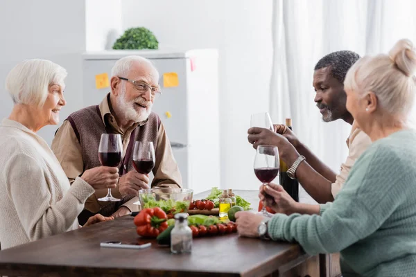 Amigos interraciales alegres y retirados sosteniendo copas con vino cerca de verduras en la mesa - foto de stock