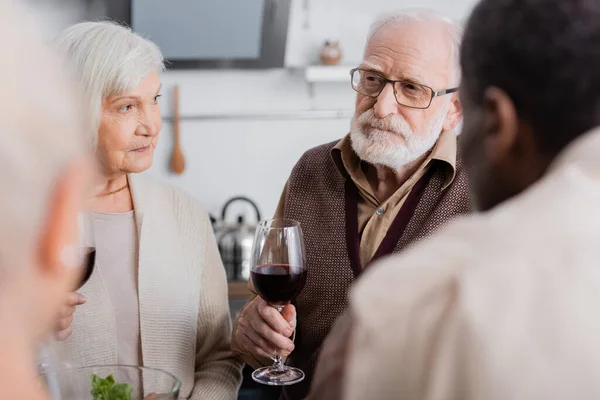 Vieil homme en lunettes tenant un verre de vin rouge près d'amis multiethniques à la retraite — Photo de stock