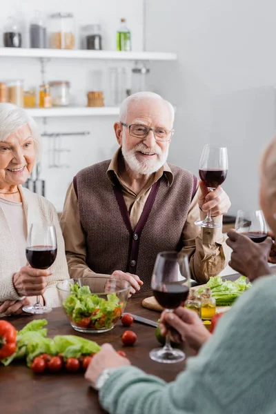 Amis interracial seniors tenant des verres avec du vin et souriant près des légumes sur la table — Photo de stock