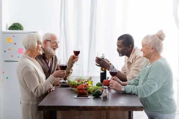 Mayores amigos interracial sosteniendo vasos con vino y sonriendo cerca de verduras en la mesa - foto de stock