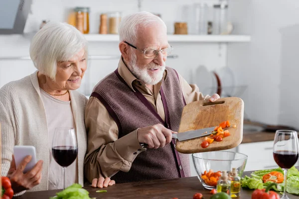 Homem sênior feliz adicionando legumes fatiados em tigela com salada perto alegre esposa aposentada com smartphone — Fotografia de Stock