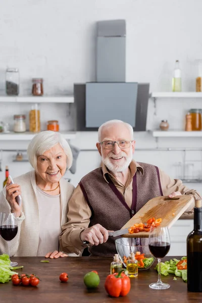 Felice uomo anziano aggiungendo verdure a fette in ciotola con insalata vicino moglie in pensione con smartphone — Foto stock