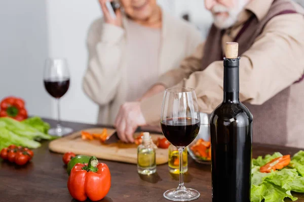 Botella con vino tinto cerca de vidrio, verduras y pareja jubilada sobre fondo borroso - foto de stock