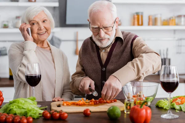 Старший мужчина режет овощи около счастливой жены на пенсии разговаривая на смартфоне — стоковое фото