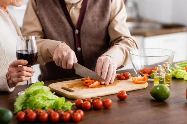 Vue recadrée d'un homme âgé coupant des légumes près d'une femme retraitée avec un verre de vin — Photo de stock