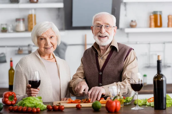 Feliz hombre mayor cortando verduras cerca de la esposa jubilada con un vaso de vino - foto de stock