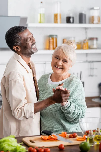 Marido y esposa multiculturales mayores sonriendo mientras se toman de la mano en la cocina - foto de stock