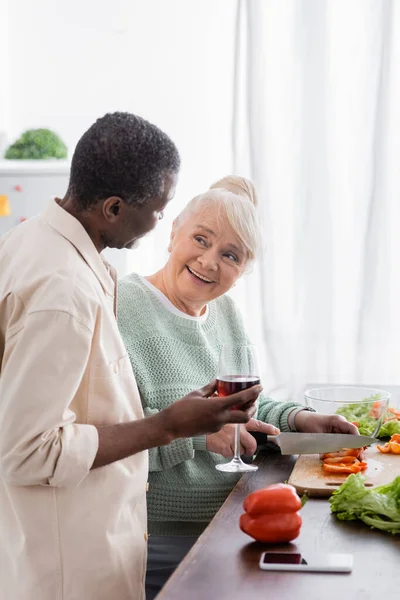 Happy senior woman cutting bell pepper near african american husband with glass of wine — Stock Photo