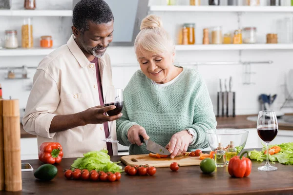 Feliz jubilado mujer corte maduro pimiento campana cerca africano americano marido con vaso de vino - foto de stock