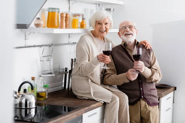 Joyeux couple âgé tenant des lunettes sur le vin rouge et regardant la caméra dans la cuisine — Photo de stock