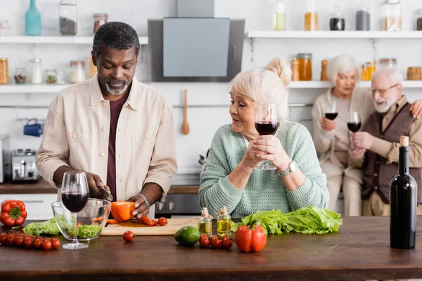 African american man cutting bell pepper near senior woman and retired friends on blurred background — Stock Photo