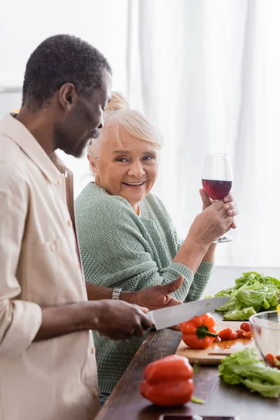 Uomo afroamericano taglio peperone vicino moglie anziana con bicchiere di vino — Foto stock