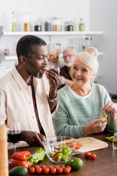 Hombre afroamericano comiendo tomate cereza cerca de la mujer mayor y amigos jubilados en un fondo borroso - foto de stock