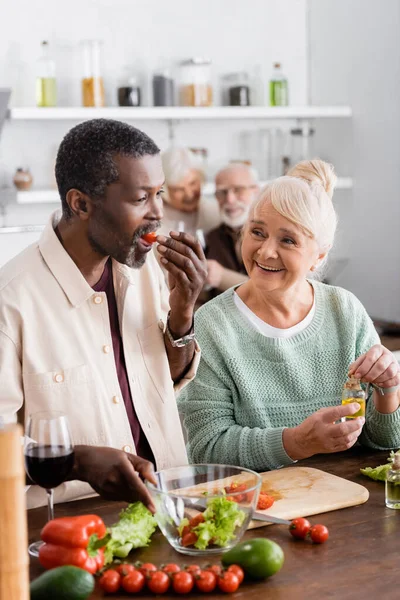 Uomo afroamericano mangiare pomodoro ciliegia vicino alla donna anziana e gli amici in pensione su sfondo sfocato — Foto stock