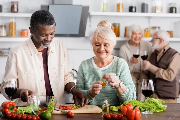 Hombre afroamericano y mujer mayor feliz preparando ensalada cerca de amigos jubilados en un fondo borroso - foto de stock