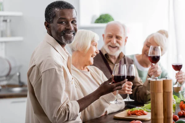 Feliz hombre afroamericano retirado sonriendo mientras sostiene una copa de vino cerca de amigos en un fondo borroso — Stock Photo