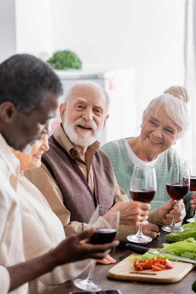 Felice uomo in pensione sorridente mentre tiene un bicchiere di vino vicino ad amici multiculturali in primo piano sfocato — Foto stock