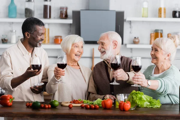 Jubilados multiculturales alegres sosteniendo copas de vino cerca de verduras en la mesa — Stock Photo