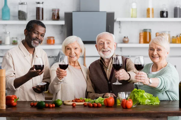 Jubilados multiculturales gozosos sosteniendo copas de vino cerca de verduras frescas en la mesa - foto de stock