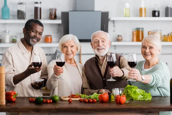 Jubilados multiculturales gozosos sosteniendo copas de vino cerca de verduras frescas en la mesa — Stock Photo