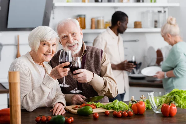 Feliz hombre y mujer de edad avanzada tintineo vasos de vino cerca de amigos multiculturales sobre fondo borroso — Stock Photo