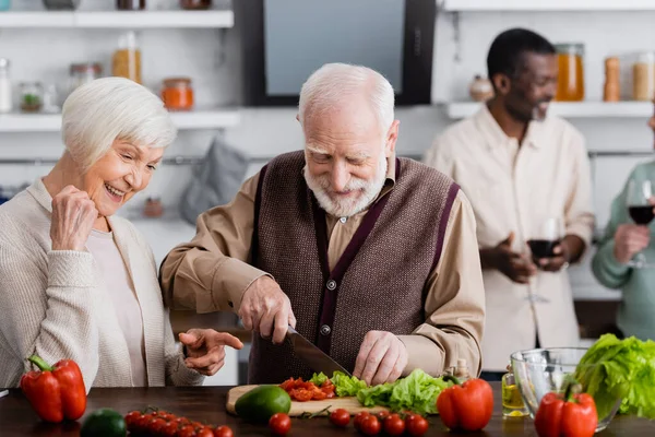 Feliz mujer mayor señalando con el dedo en el hombre jubilado ensalada de cocina cerca de amigos multiculturales sobre fondo borroso - foto de stock