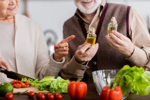 Ausgeschnittene Ansicht einer glücklichen Seniorin, die mit dem Finger auf Ölflaschen in der Nähe eines Rentners zeigt — Stockfoto