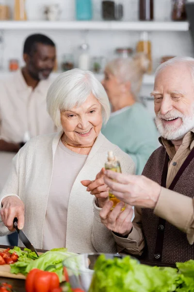 Feliz mujer mayor señalando con el dedo en la botella con aceite cerca de hombre jubilado y amigos multiculturales sobre fondo borroso - foto de stock