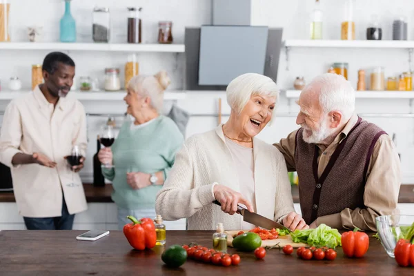 Щасливий старший чоловік і жінка готують салат біля багатоетнічних друзів на розмитому фоні — стокове фото