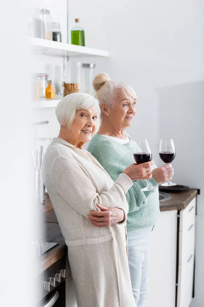 Mujeres jubiladas felices sosteniendo vasos con vino en la cocina - foto de stock